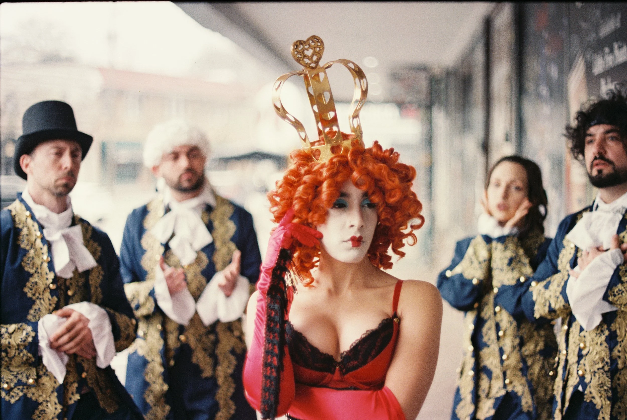 A woman with bright red, curly hair wearing a red and black lace slip dress, leading a group of men dressed in ornate costumes, in what appears to be a theatrical or performance setting.