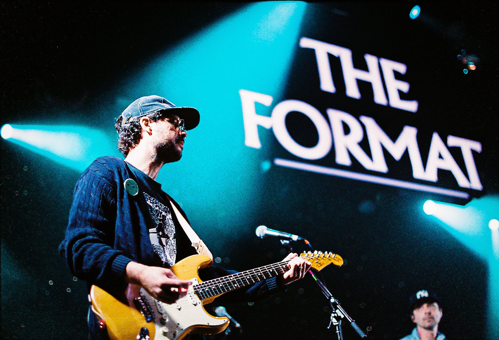 A musician playing an electric guitar on stage with a blue spotlight shining behind him. He is wearing a dark jacket, a hat, and glasses, with a large button pinned to his chest. In the background, there's a neon sign that reads 'THE FORMAL' and a partially visible band member.