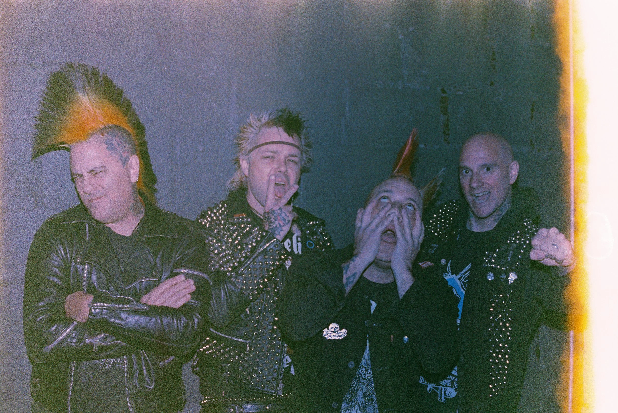 Four individuals posing against a gray wall, wearing punk-style clothing with spikes and leather, two of them with mohawk hairstyles, one with a devil's tail tattoo, and expressive facial expressions.