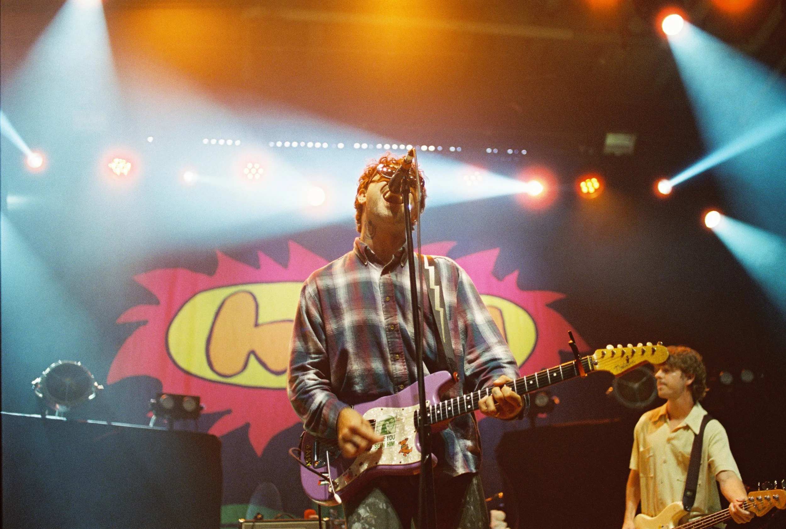 A musician with curly red hair, wearing glasses and a plaid shirt, performs on stage with a guitar and a microphone. Stage lights illuminate the scene, and a colorful backdrop with the word 'Coco' can be seen behind him. Other band members are partially visible, and a young girl stands nearby.