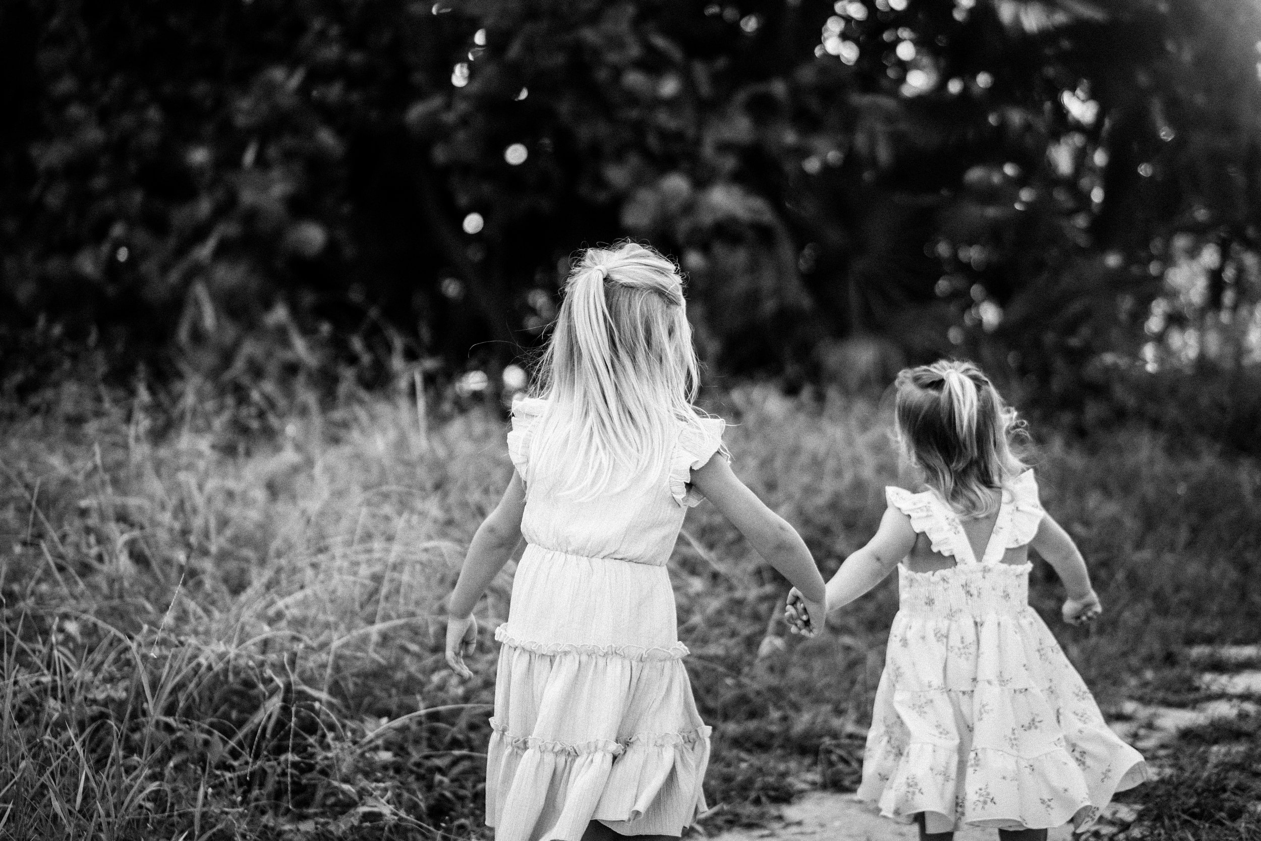 Two young girls holding hands and walking in a grassy outdoor area with trees in the background. The image is in black and white.
