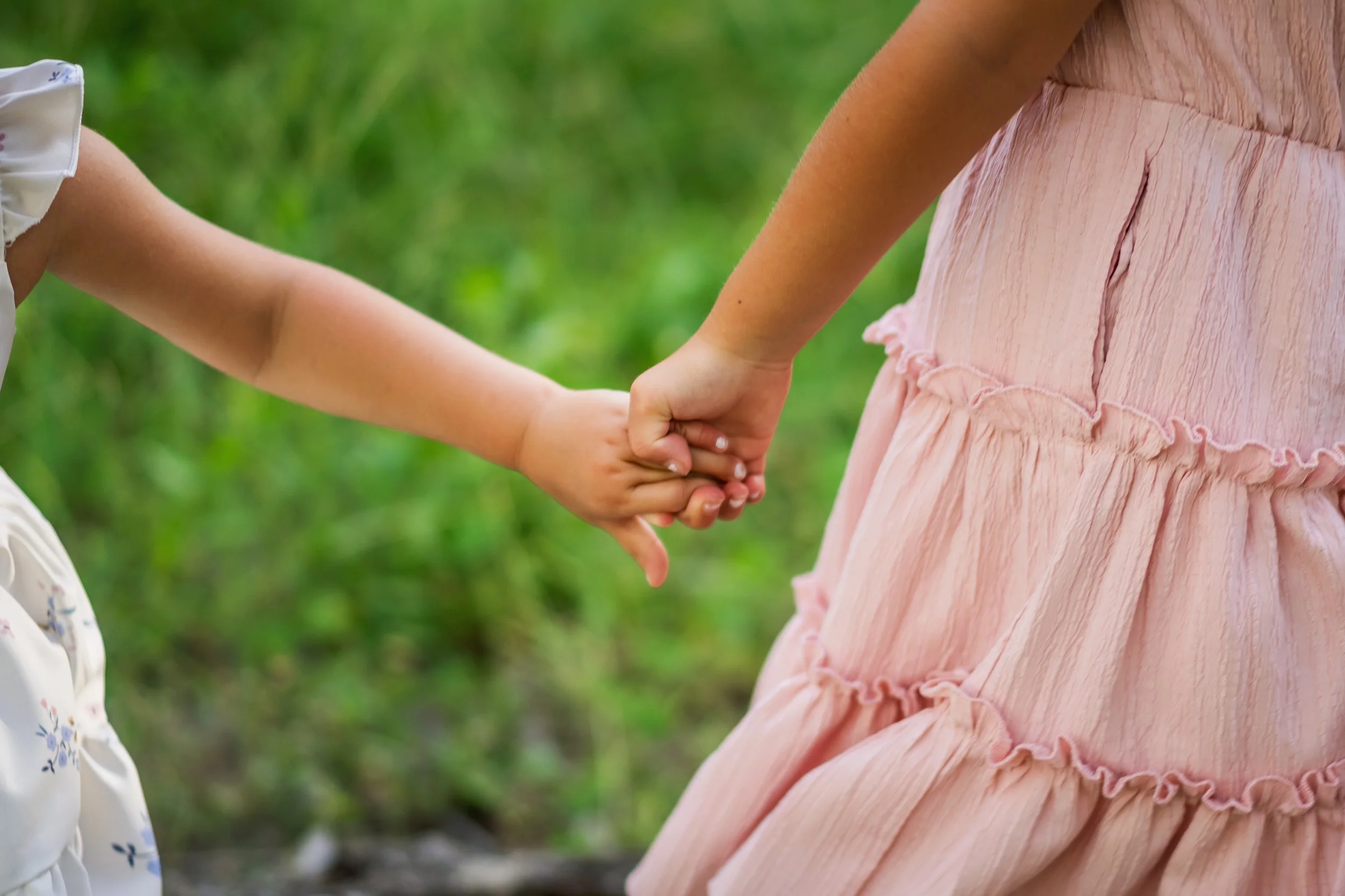Two young girls holding hands outdoors with greenery in the background, one wearing a pink dress and the other in a white dress with a floral pattern.