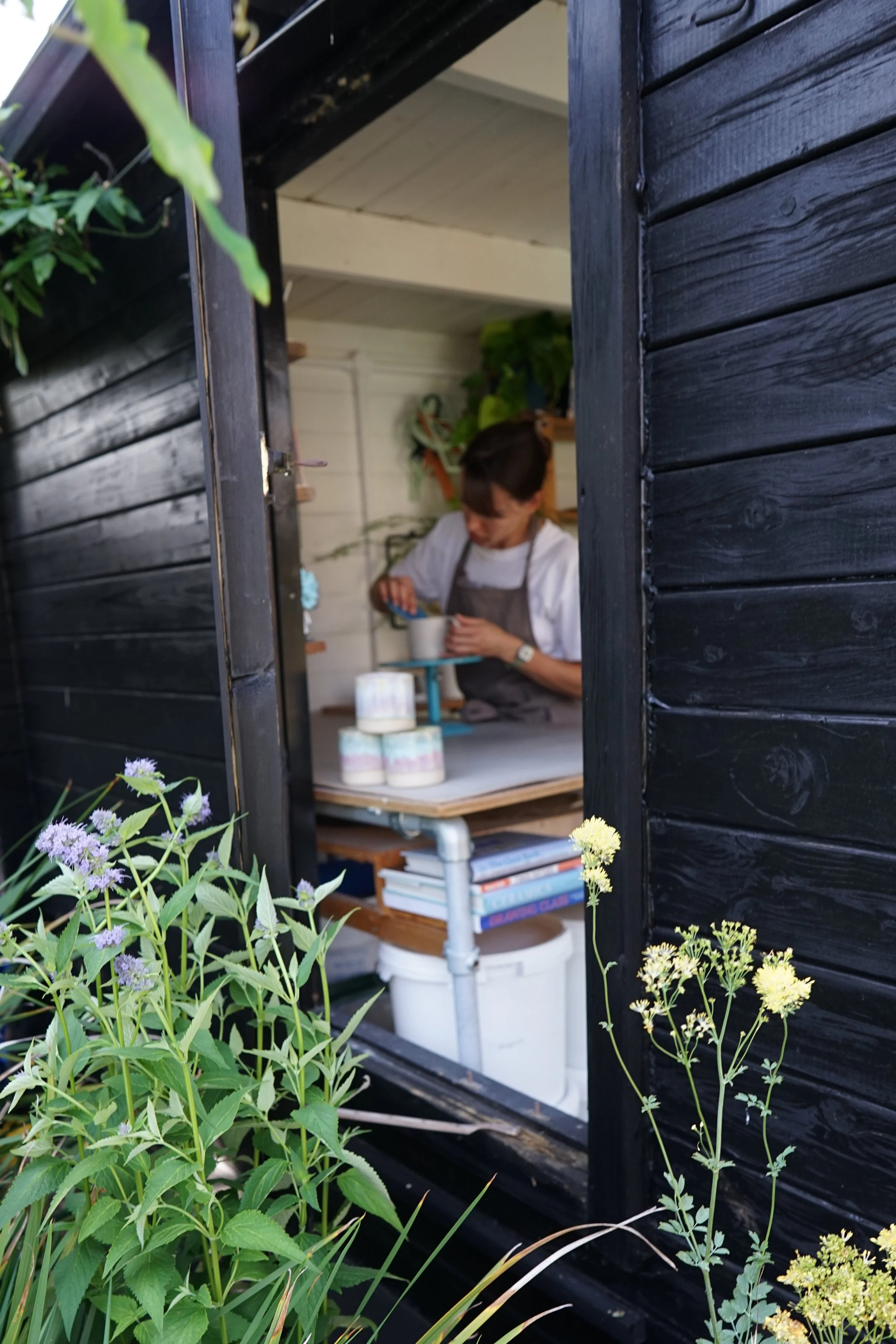 View through a window showing a person working on a craft project inside a room, with plants and shelves visible, framed by black wooden siding and surrounded by green plants and flowers outside.