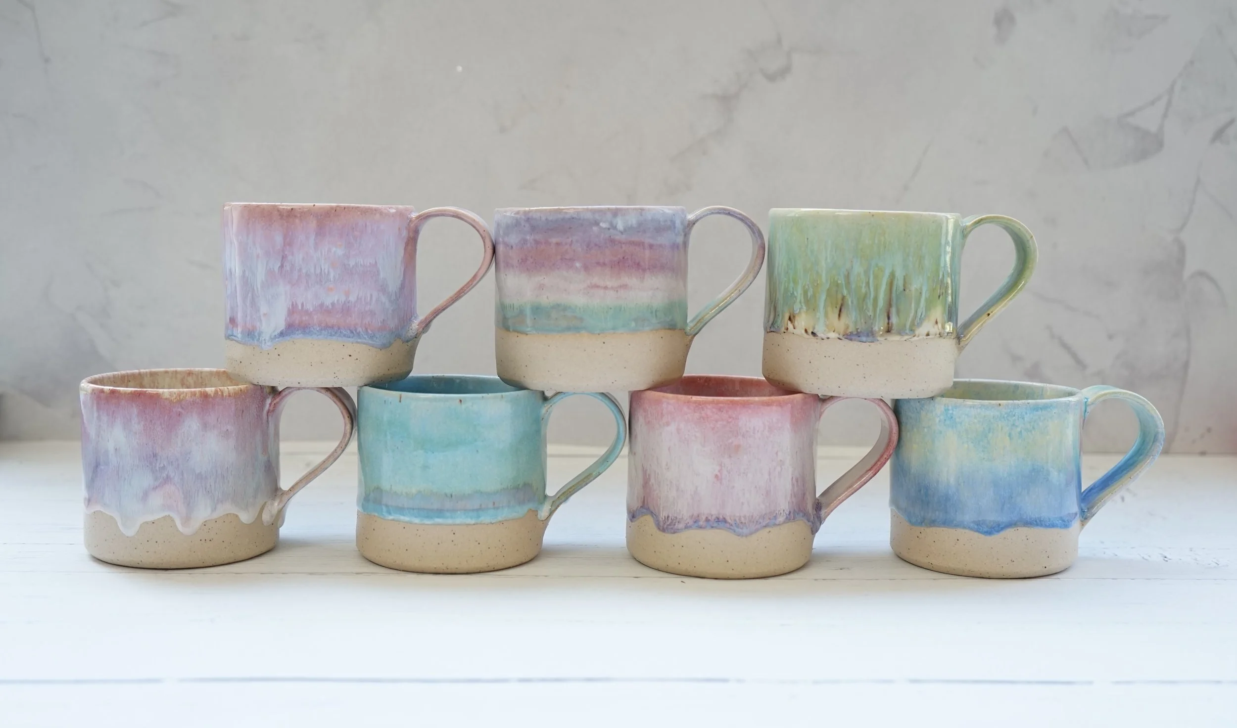 Stacked colorful ceramic mugs with pastel glaze on a white surface against a textured light gray background.