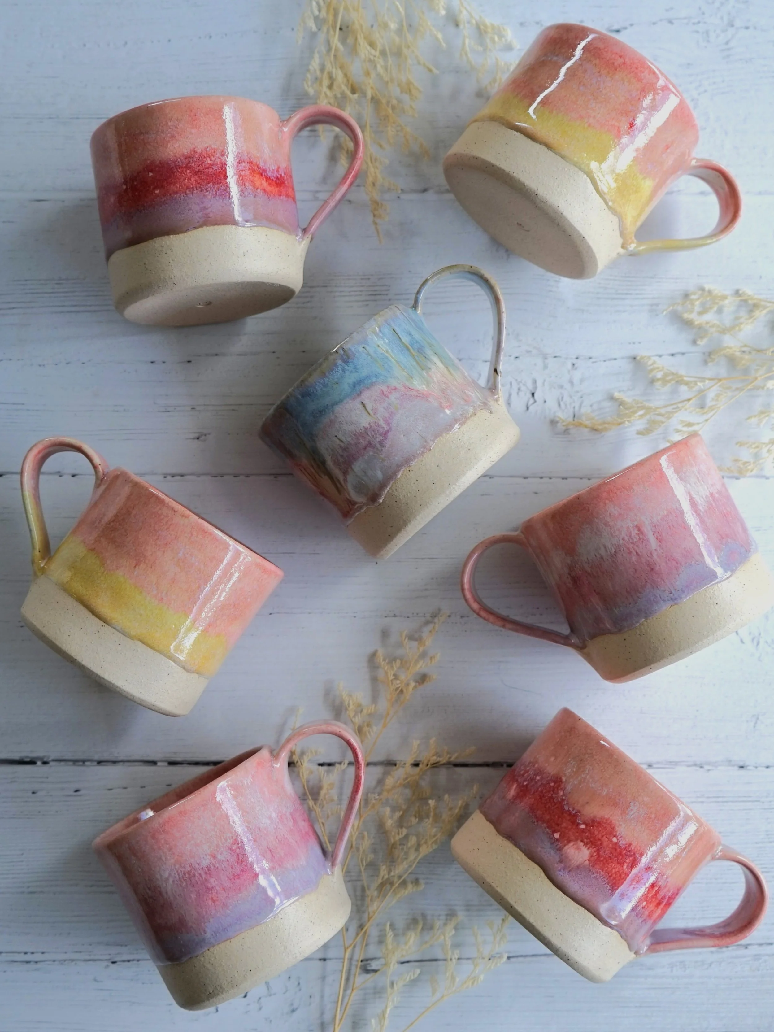 Six ceramic mugs with pastel-colored abstract glaze designs arranged on a white wooden surface with dried plant decorations.