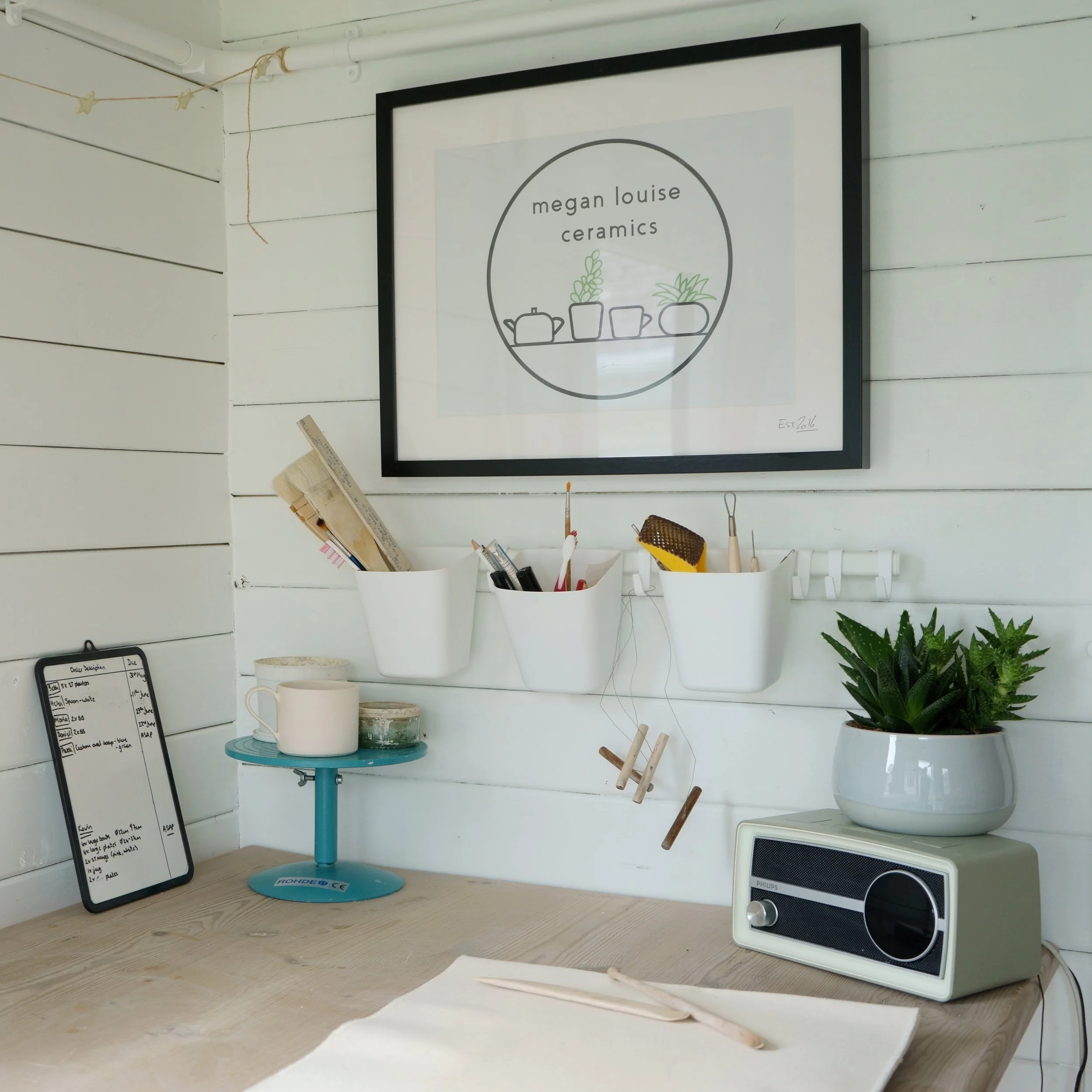 A workspace with a framed sign reading 'Megan Louise Ceramics' and illustrations of potted plants. The workspace has a white wood wall with a string of star-shaped lights, a small blue table with a white mug, and a variety of tools and potted plants on the desk.