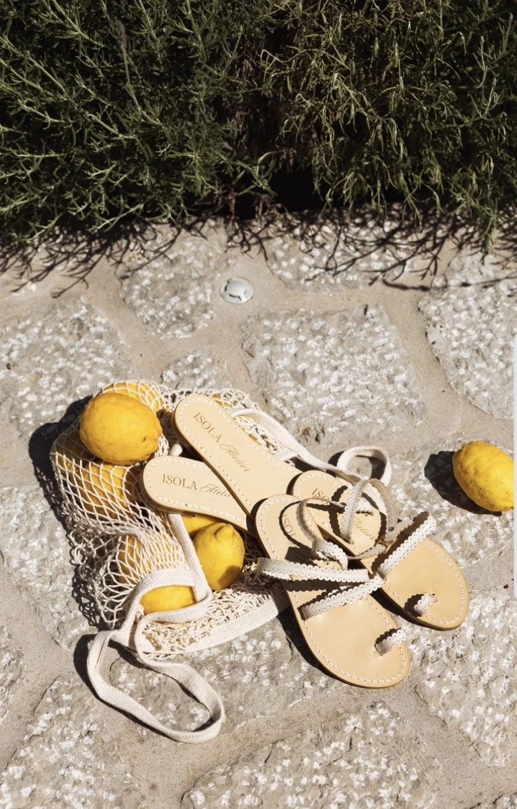 A pair of beige sandals with black trim, placed on a stone-paved surface, surrounded by lemons and a white string bag. Green shrubbery is visible in the background.
