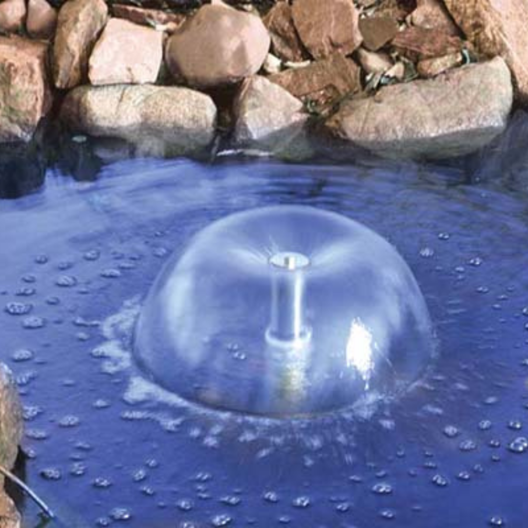 A large, transparent, dome-shaped water fountain in a pond surrounded by rocks.