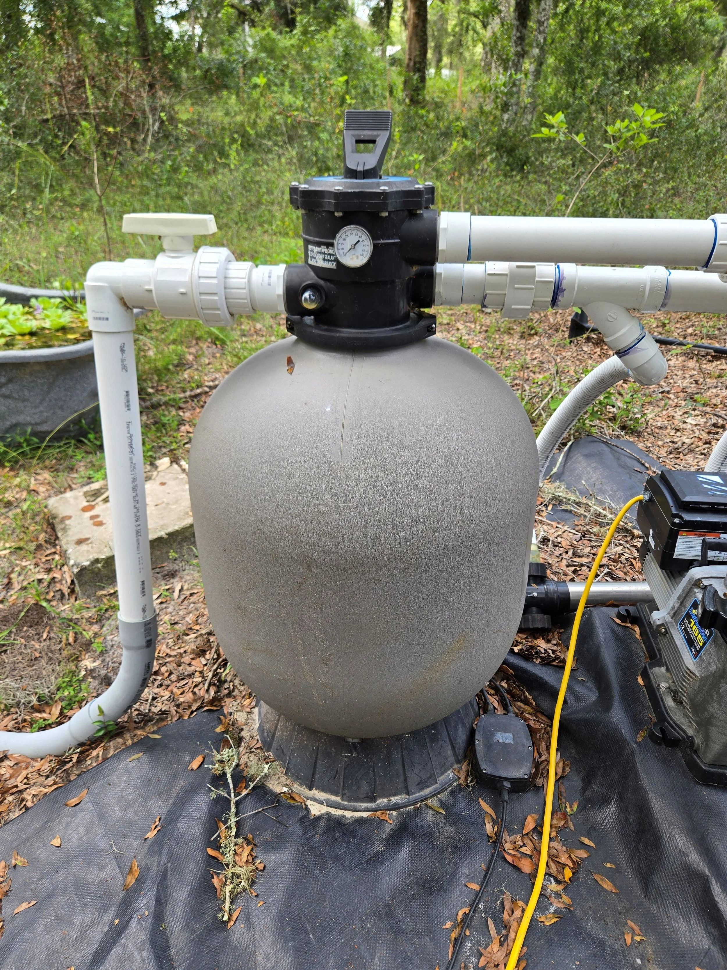 A water filtration system with a large grey tank, connected to white PVC pipes, is set up outdoors amidst trees and fallen leaves on black ground cover.
