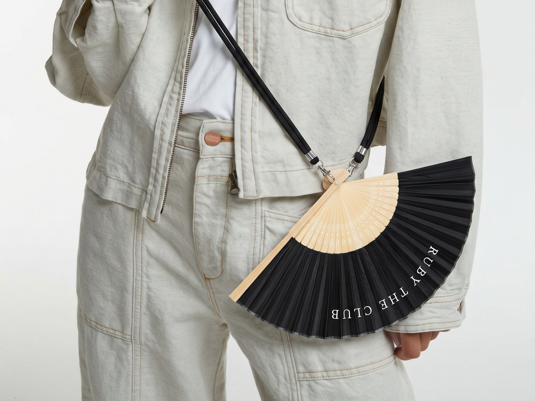 Person wearing beige denim jacket and pants, holding a black and beige folding fan with the words "THE RUBY CLUB" printed on it.