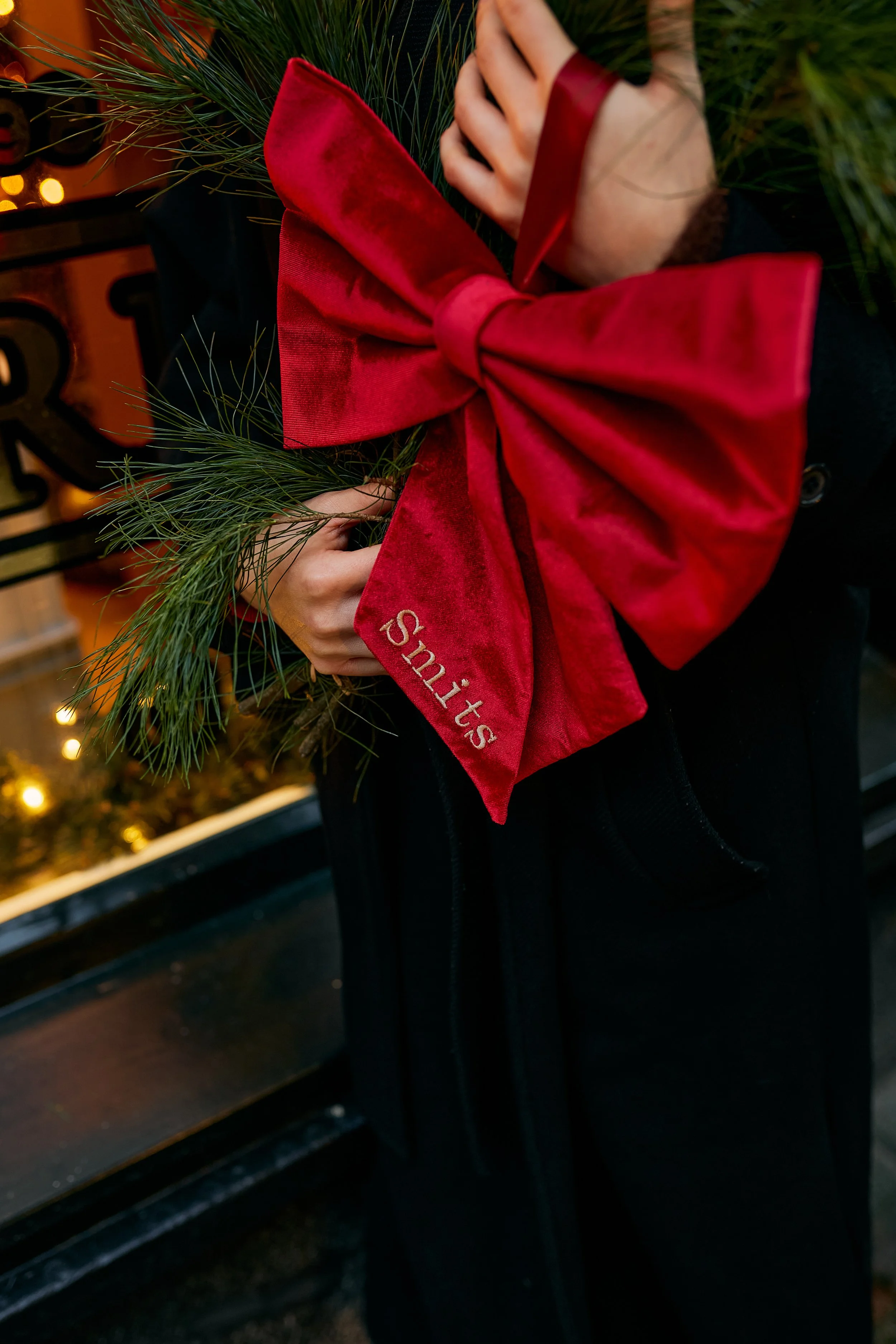 A person holding a small Christmas wreath decorated with red velvet ribbon and pine branches in front of a black metal gate with Christmas lights in the background.