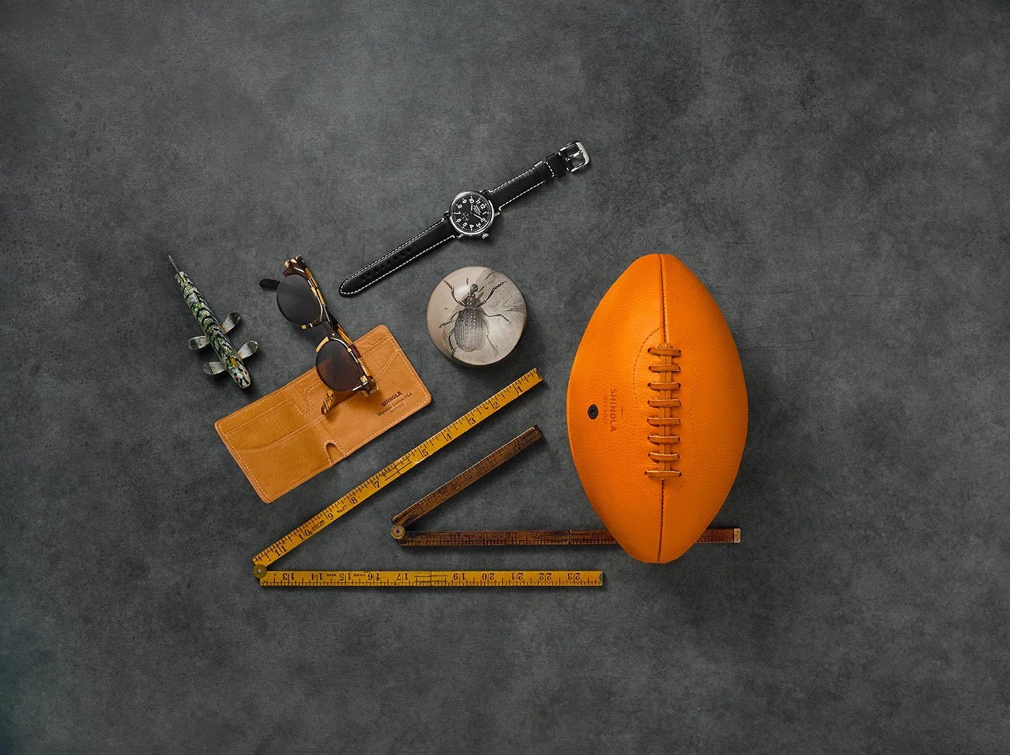 Kiyume campaign still life with a leather football, watch, sunglasses, wallet and folding rulers on a gray surface.
