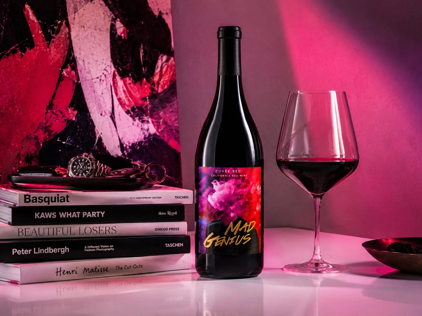 Mad Genius wine bottle and glass beside art books and abstract artwork in a pink-lit setting