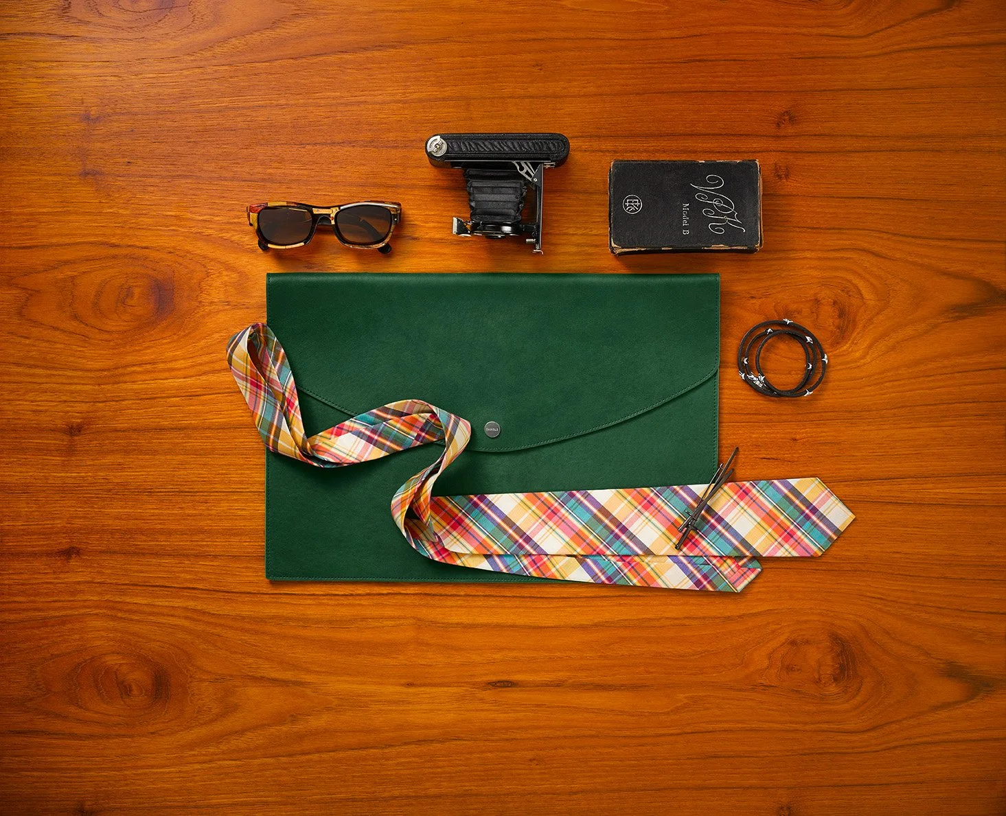 Kiyume campaign still life with a green leather folio, plaid tie, vintage camera, sunglasses and bracelets on wood.