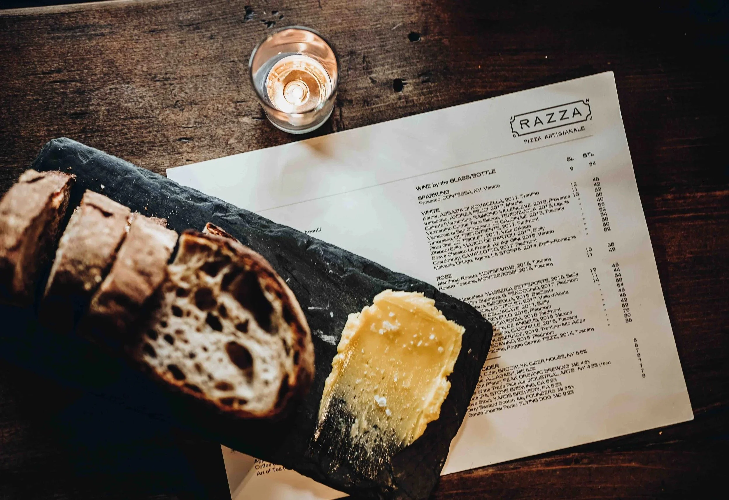 Razza wine menu on a wooden table beside sliced bread, butter and a candle.