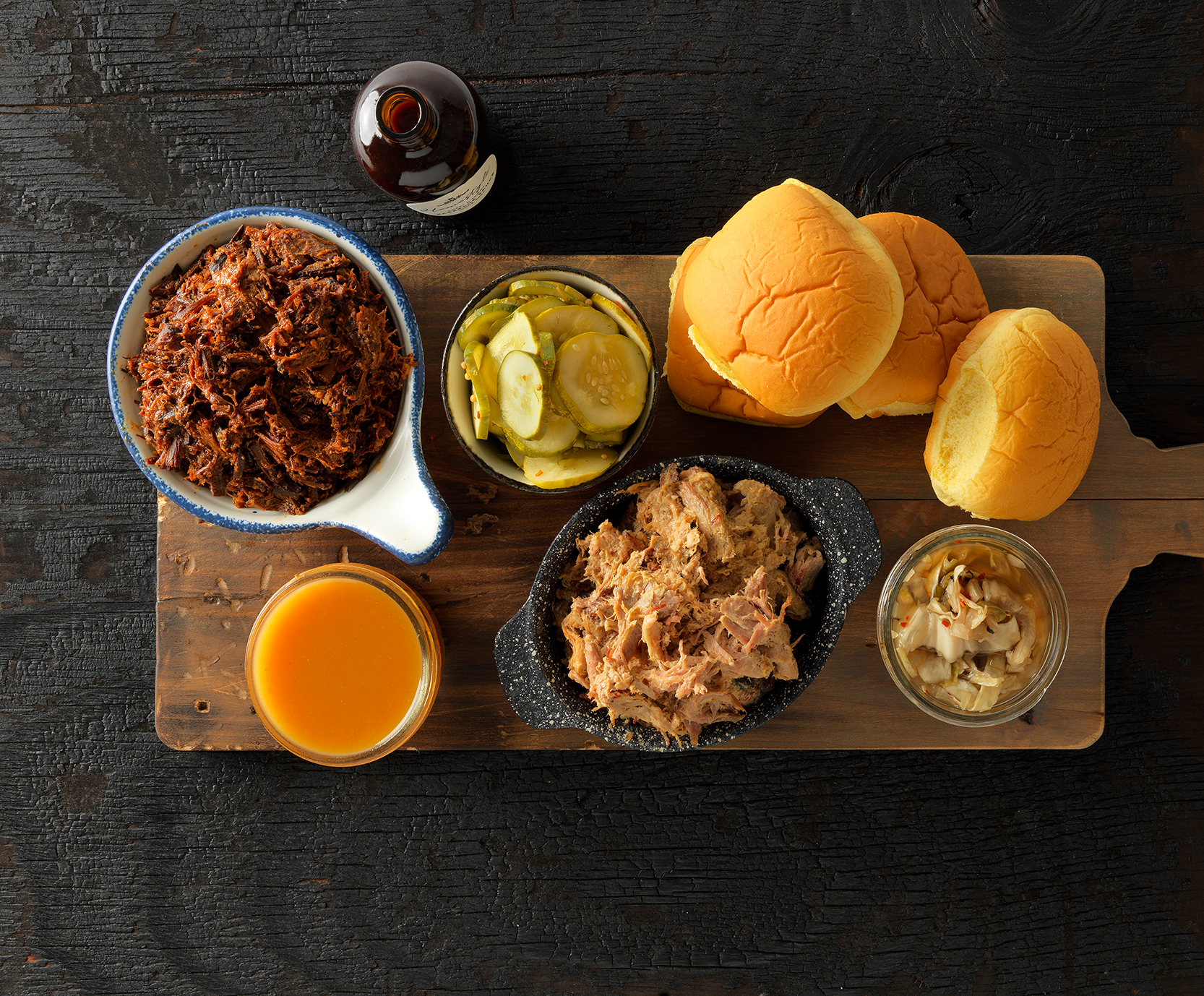 Hill Country barbecue sandwich ingredients arranged with buns, pickles, sauce and shredded meat on a wooden board.
