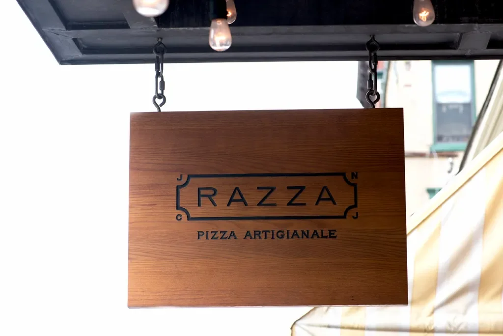Wooden hanging sign bearing the Razza Pizza Artigianale logo beneath exposed light bulbs.