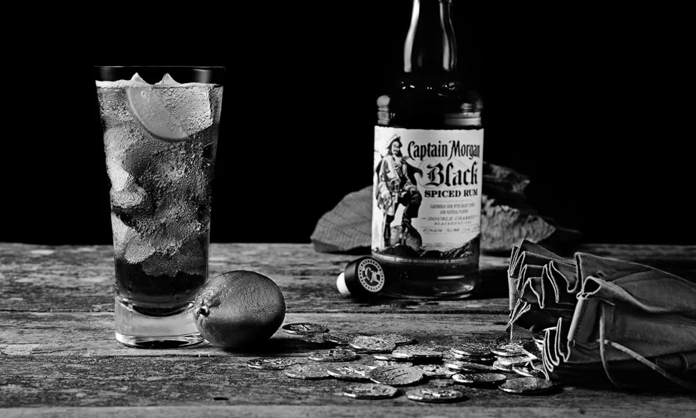 Black-and-white Captain Morgan Black bottle and tall cocktail beside a lime, coins and a drawstring pouch.