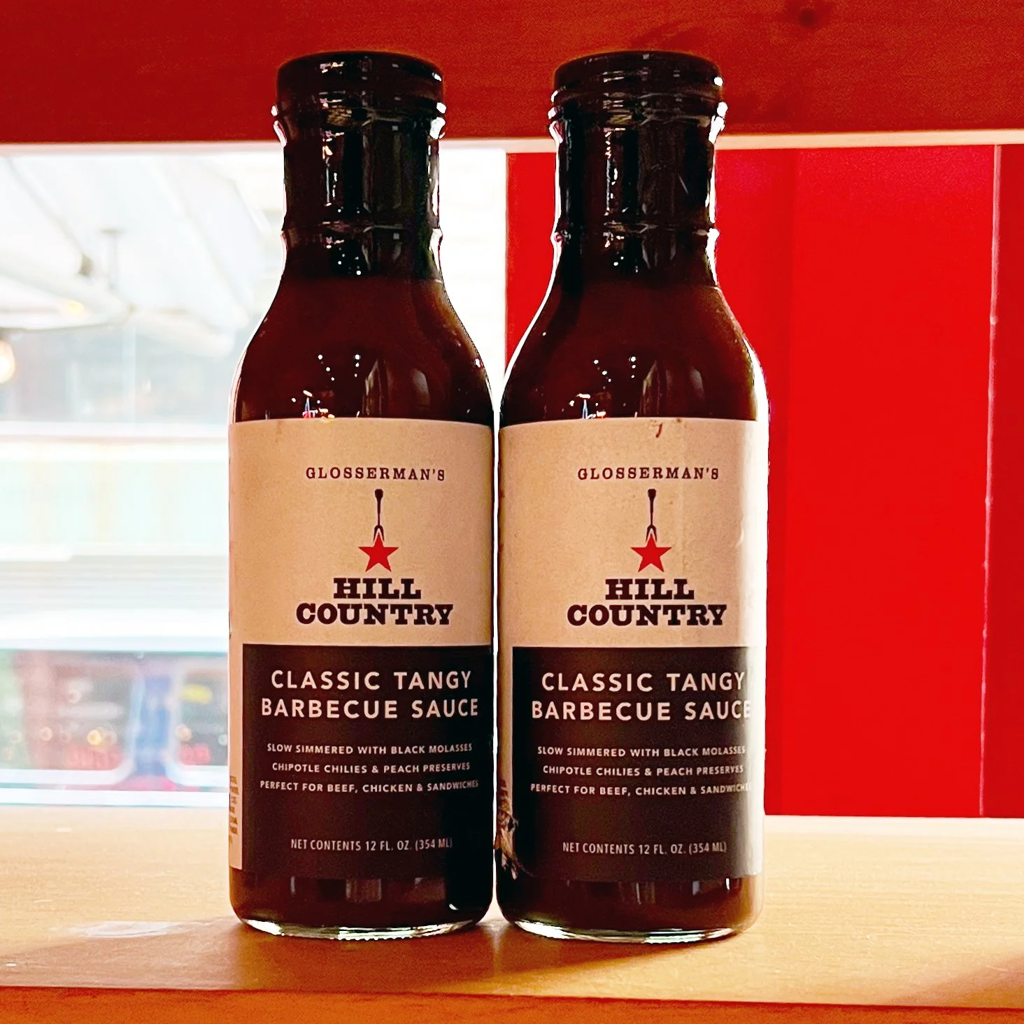 Two finished Hill Country Classic Tangy Barbecue Sauce bottles on a shelf against a red background.