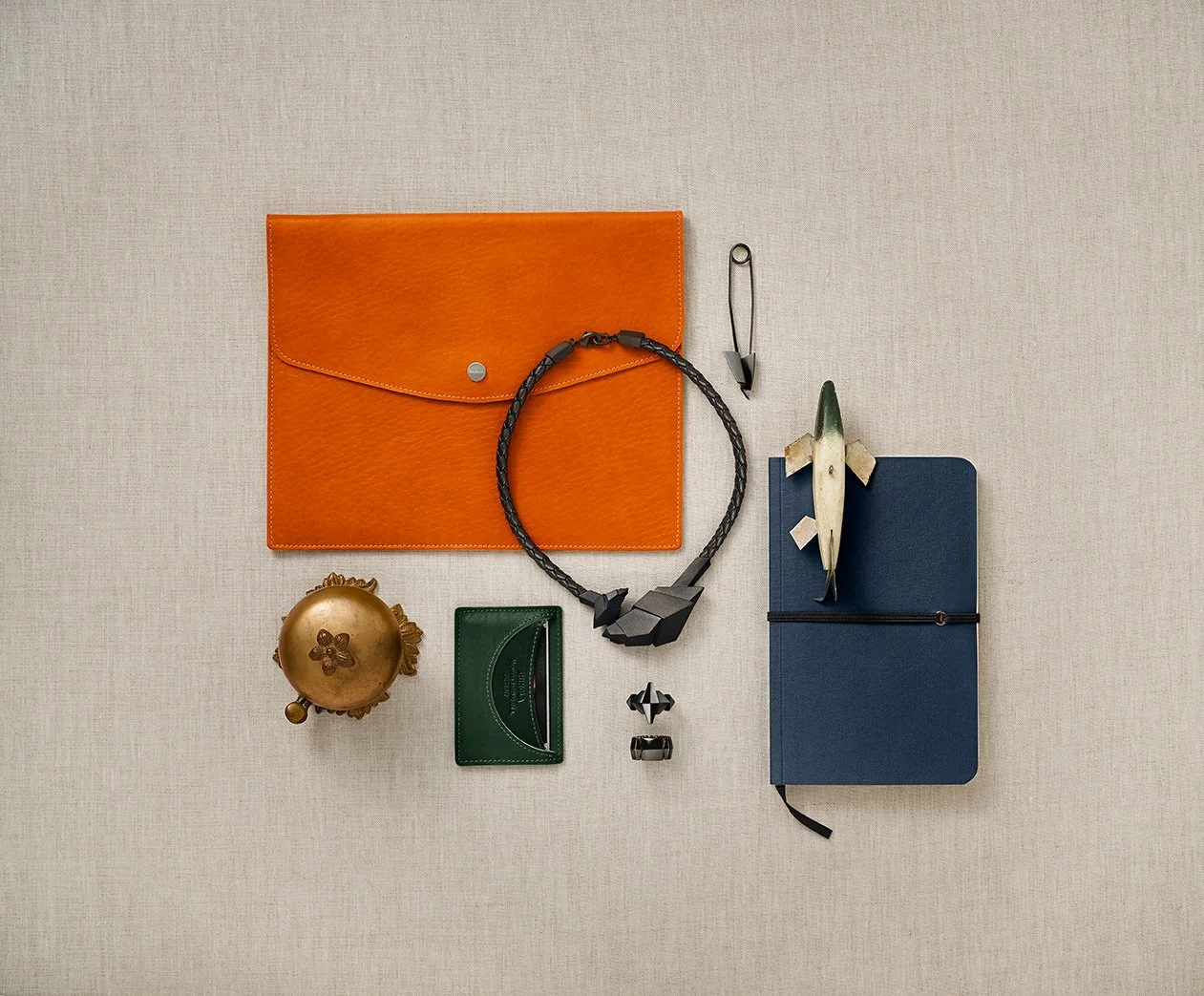 Kiyume campaign still life with an orange leather folio, angular jewelry, green wallet and navy notebook on linen.