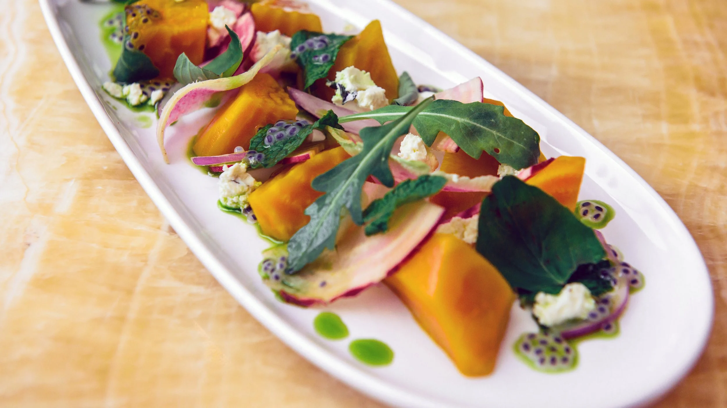 Colorful vegetable dish with leafy garnishes, cheese and sauce arranged on an oval white plate at Measure.
