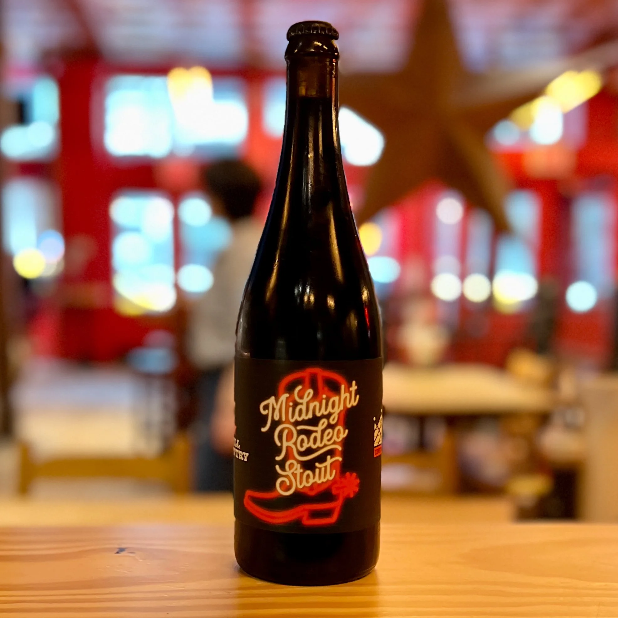 Midnight Rodeo Stout bottle on a wooden counter with the restaurant interior softly blurred behind it.