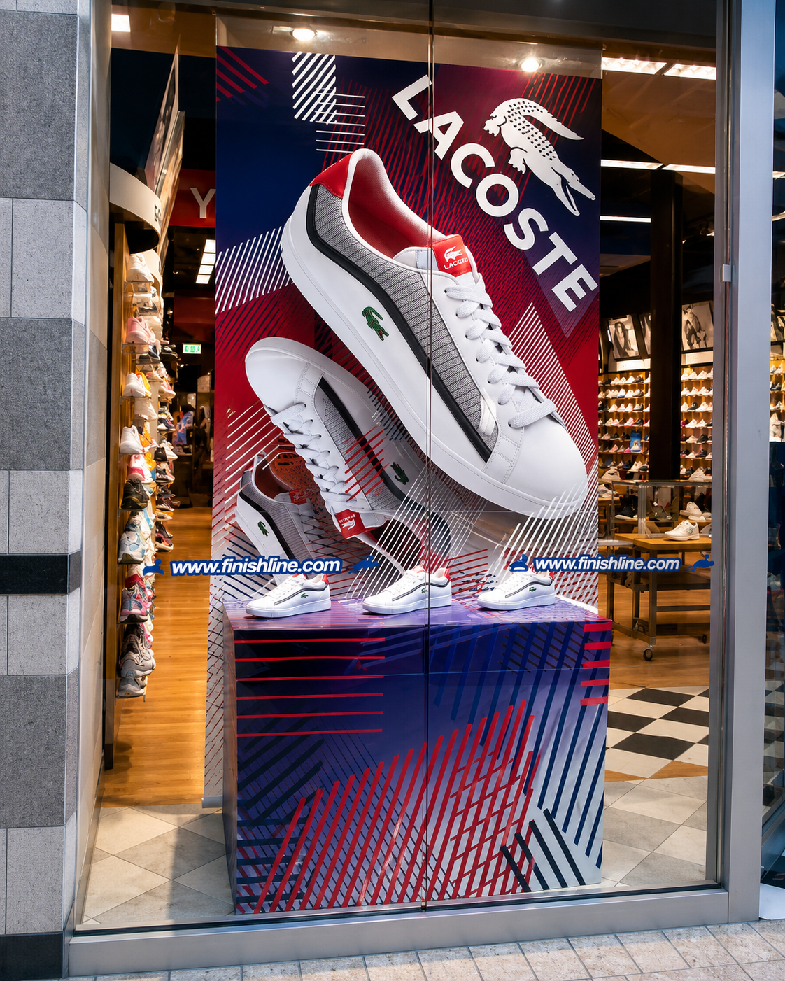 Lacoste Finish Line window display combining white sneaker photography with a red and navy patterned pedestal.