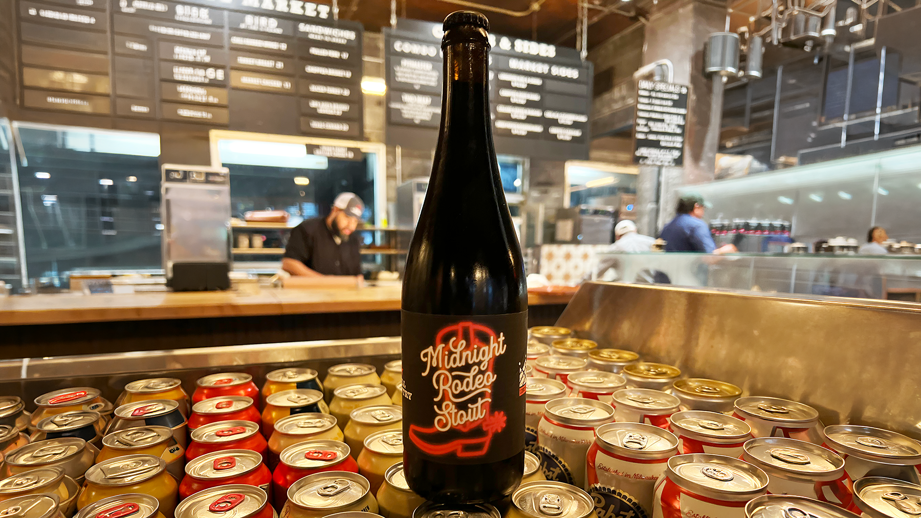 Midnight Rodeo Stout bottle displayed among beer cans in front of a restaurant counter and menu boards.