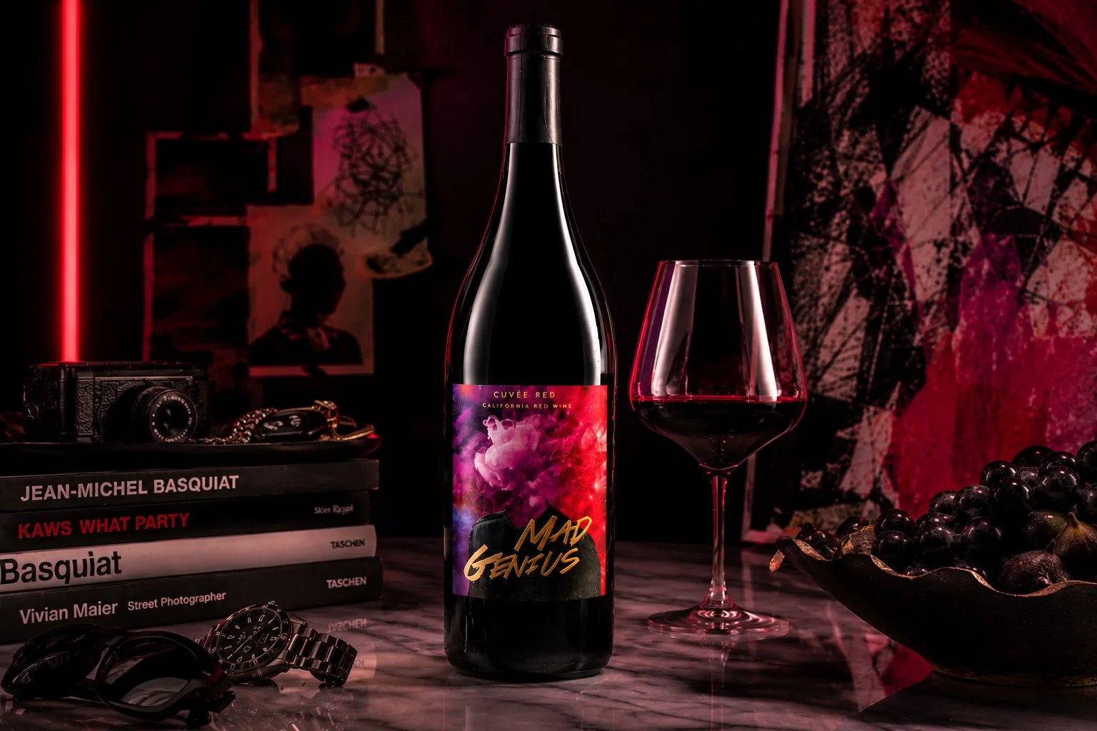 Mad Genius wine bottle and glass styled with art books, a camera and artwork in a red-lit setting