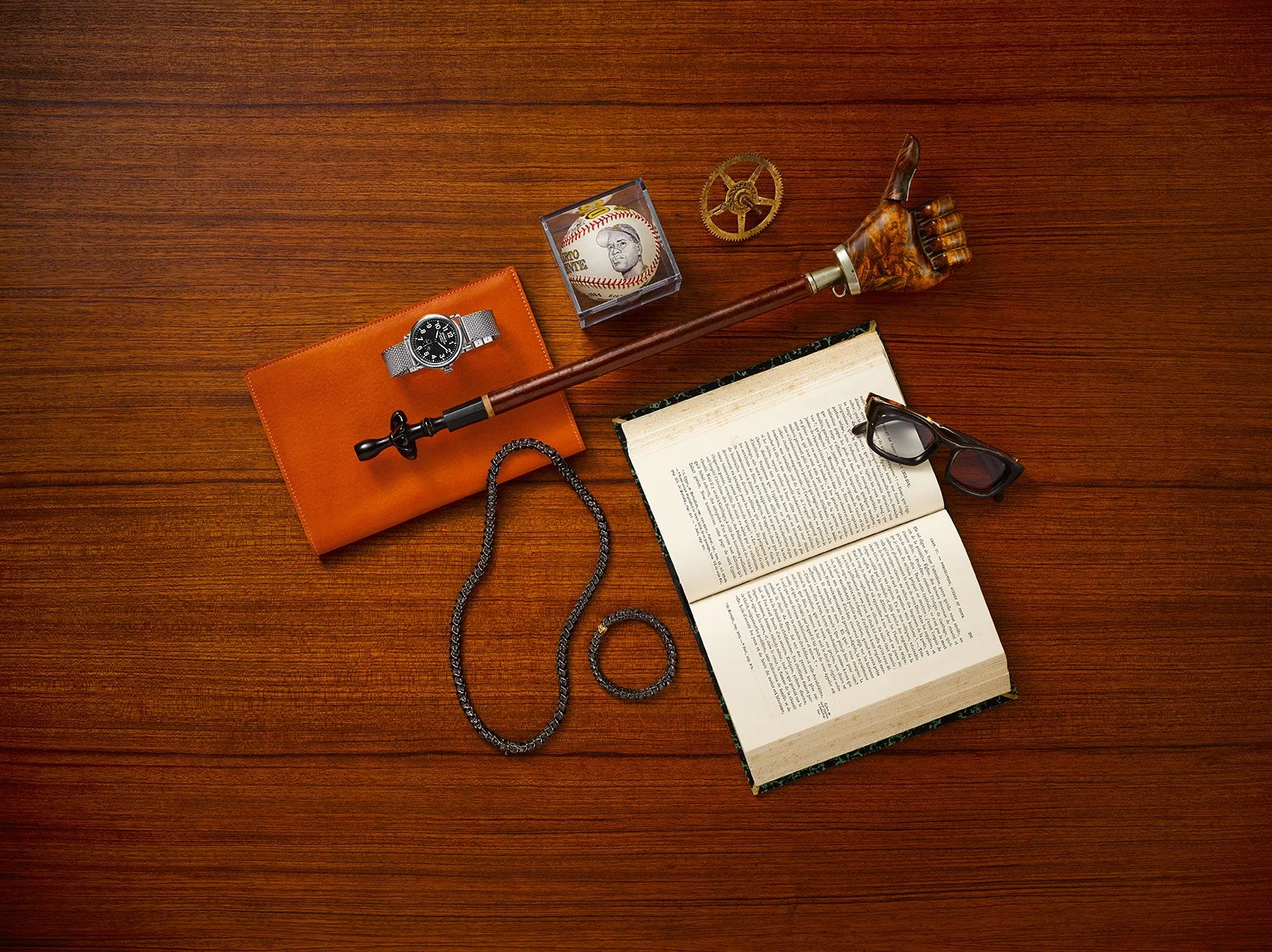 Kiyume campaign still life with a watch, leather folio, carved walking stick, jewelry and an open book on wood.