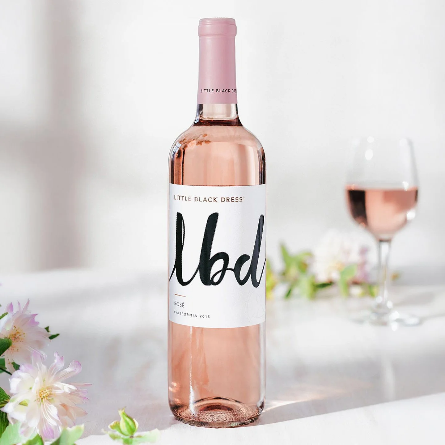 Little Black Dress rosé bottle with a white label, black script logo and pink capsule beside flowers and a wine glass.