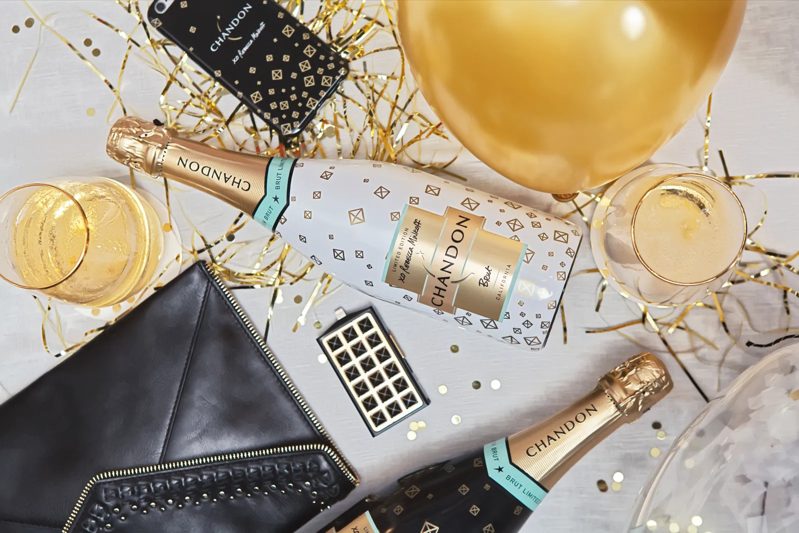Flat lay of a white Chandon × Rebecca Minkoff bottle with wine glasses, gold party decorations and accessories.