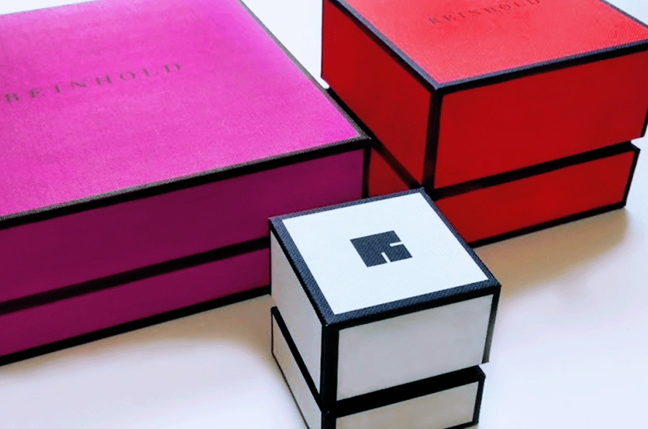 Reinhold Jewelers packaging collection with white, magenta and orange jewelry boxes outlined in black.