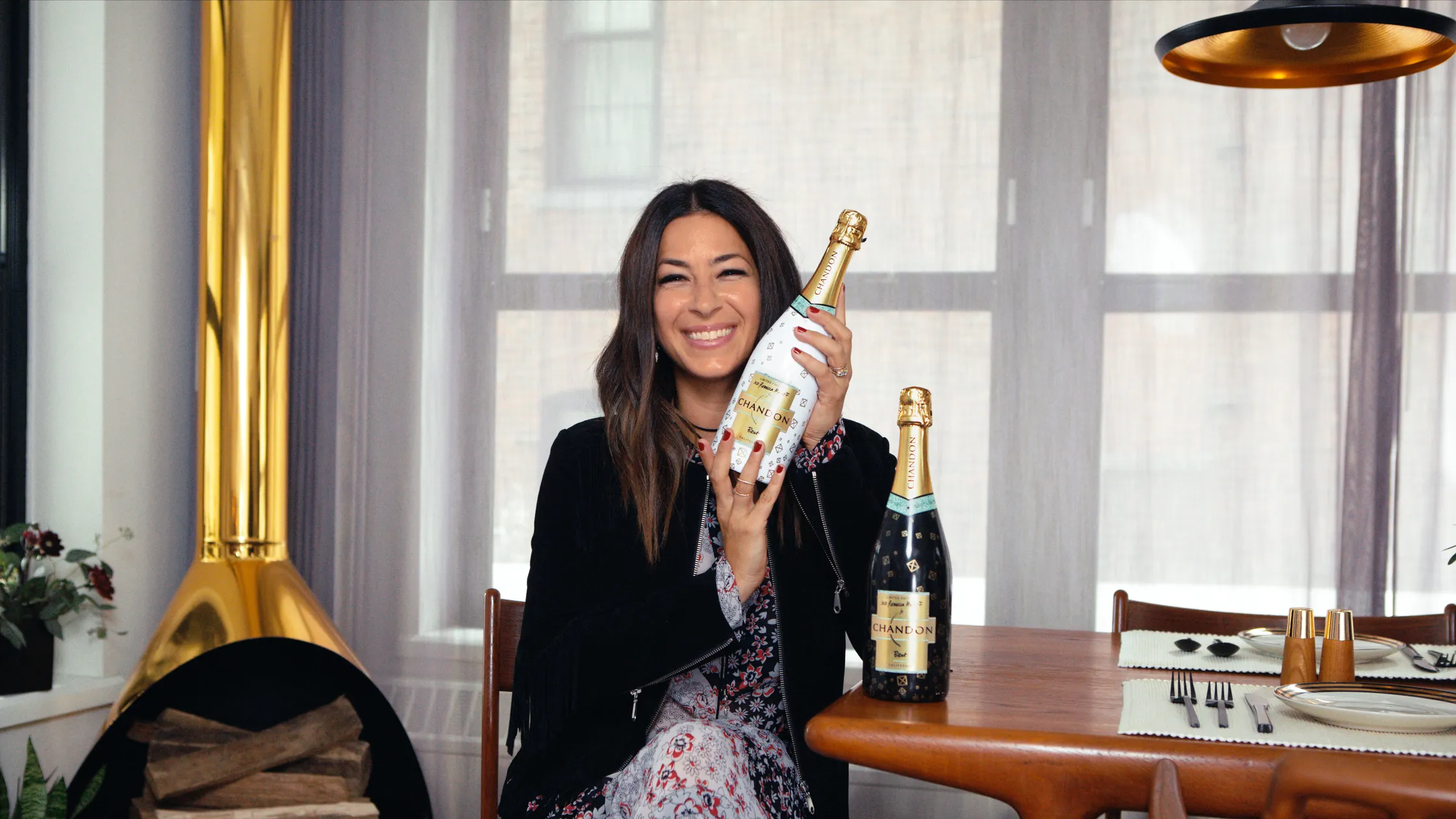 Woman holding a white Chandon × Rebecca Minkoff bottle, with a black bottle on the table beside her.