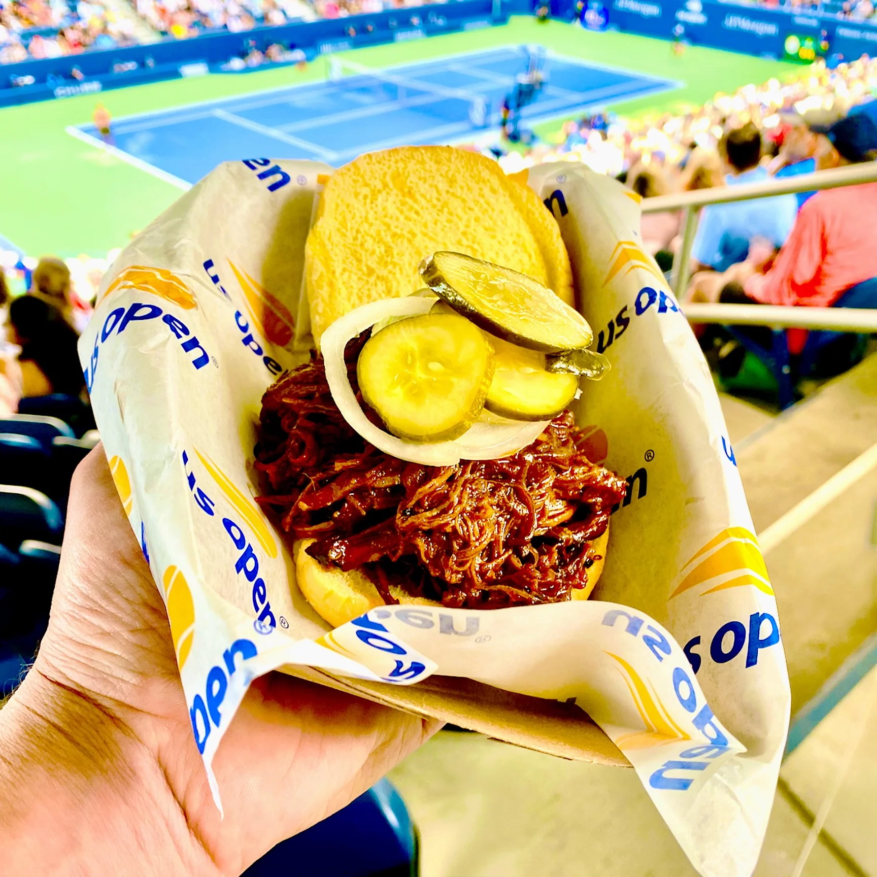 Hill Country pulled-pork sandwich with pickles held above a tennis court at the US Open.