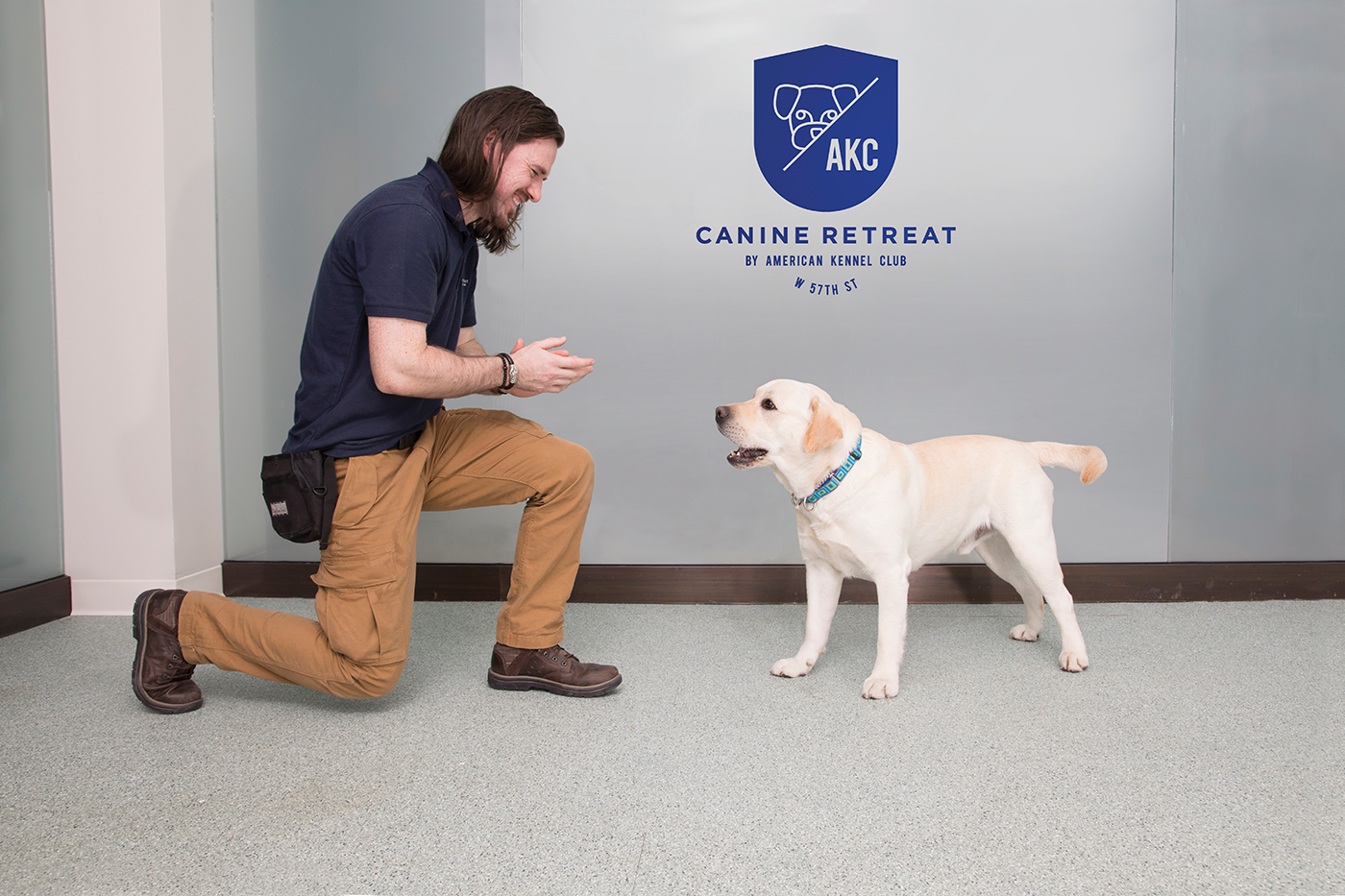 AKC Canine Retreat team member kneeling beside a dog beneath the wall-mounted logo.