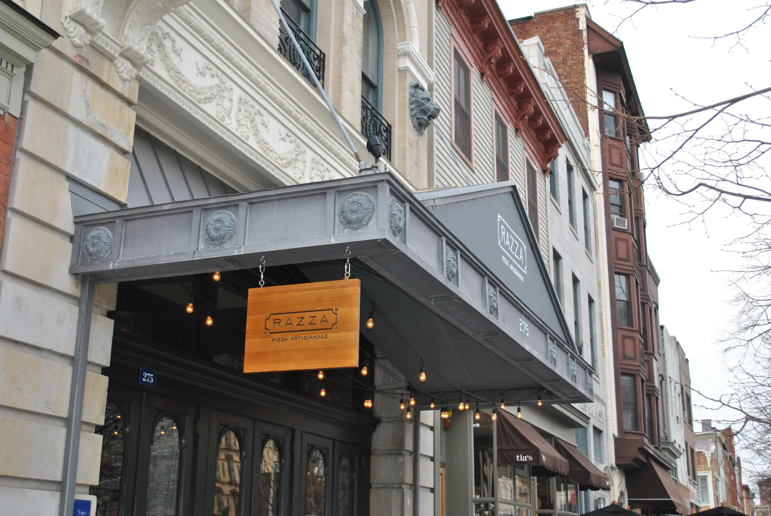 Razza restaurant exterior with branded gray awnings, a hanging wooden sign and string lights.
