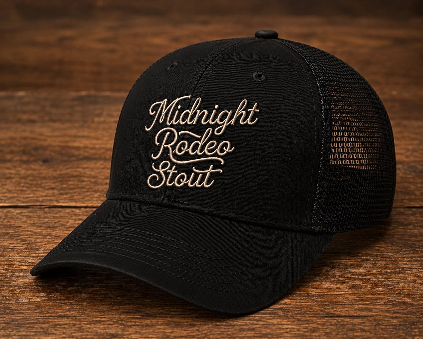 Black Midnight Rodeo Stout trucker cap with cream script embroidery on a wooden surface.