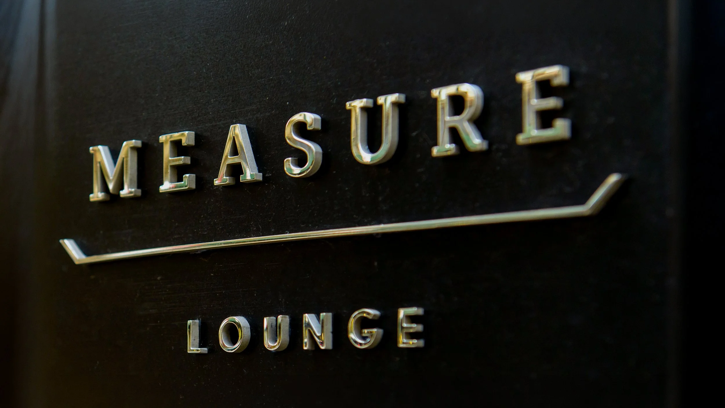 Dimensional brass Measure Lounge lettering mounted on a dark wall.