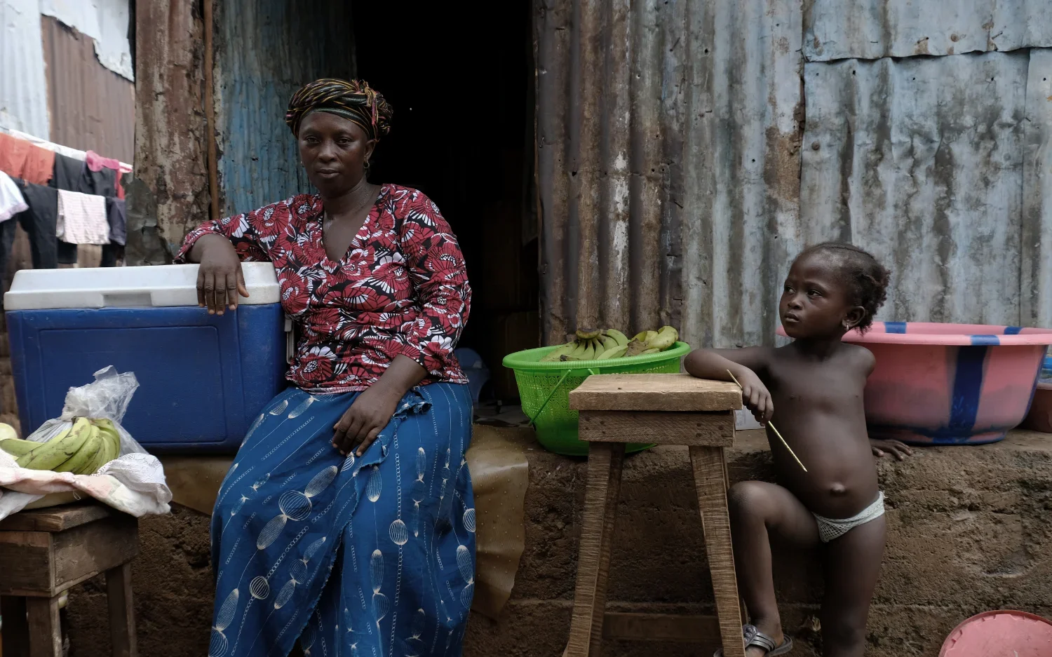 Fatamate Jalloh sits next to her streetside business in the community of Kroo Bay while a local child stands next to her people watching. A mother of seven children, Fatamate's husband died from Ebola and afterward she created her business to support