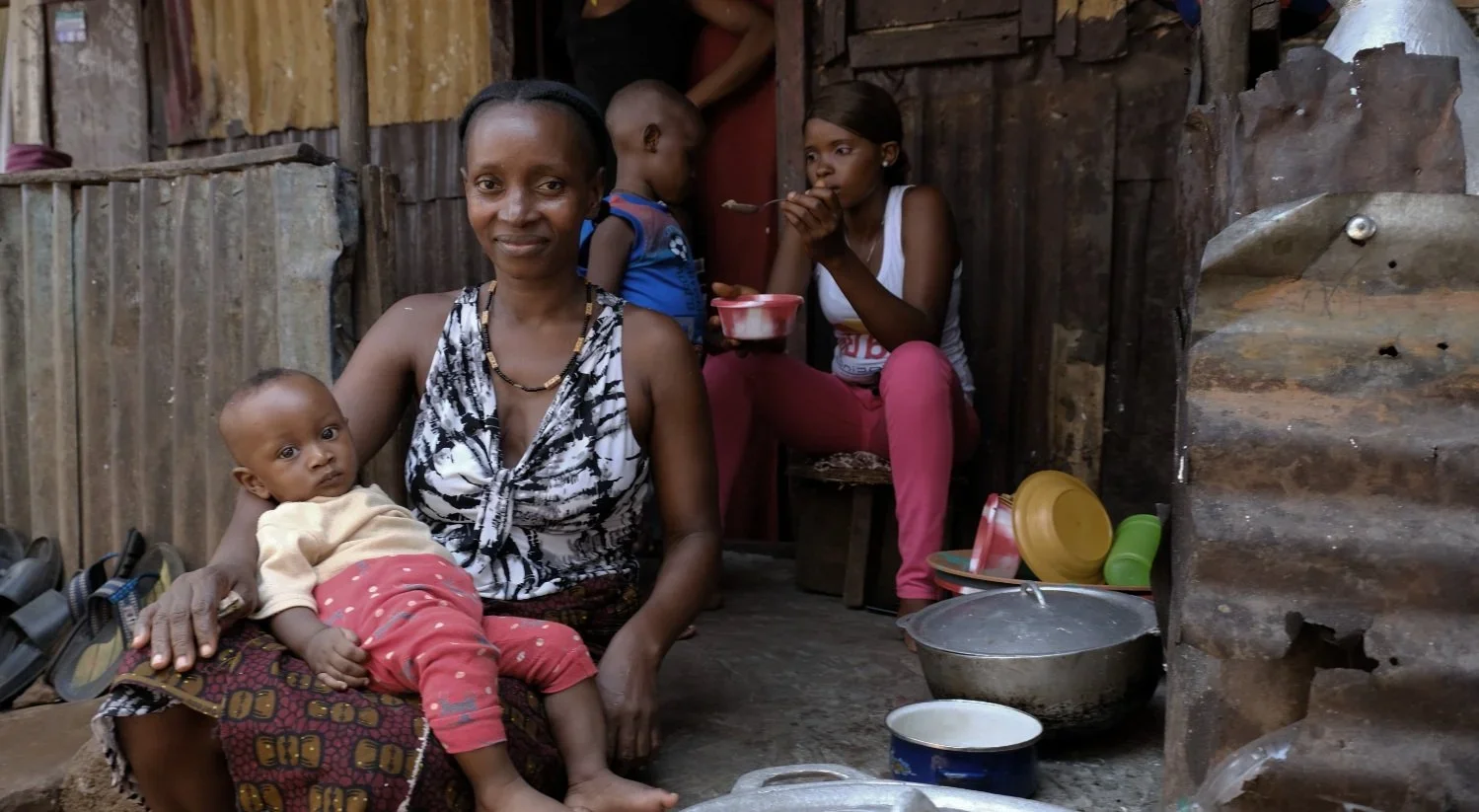 Fatemate Kamara, 41,  sits with her 6-month-old son while her daughter behind feeds her 3-year-old. Fatemate has five biological children and cares for three additional kids. One whom she adopted into her home after finding her orphaned on the street