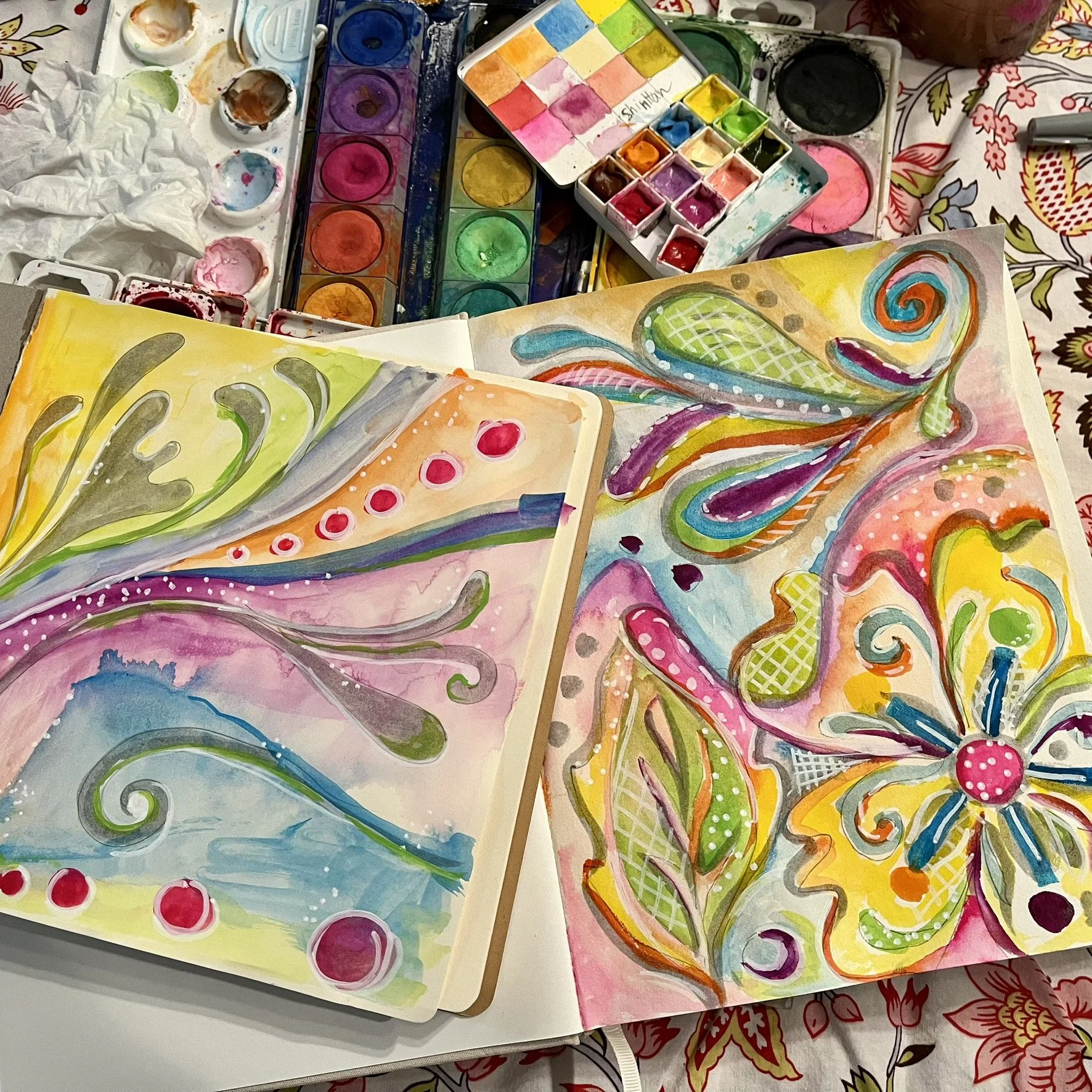 Colorful watercolor paintings of abstract floral and swirl patterns on paper, surrounded by watercolor palettes and art supplies.