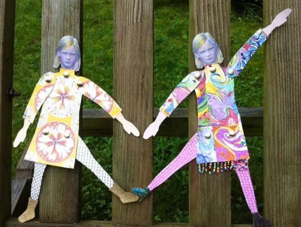 Colorful paper cutouts of two girls with painted faces, holding hands and standing between wooden fence posts outdoors.