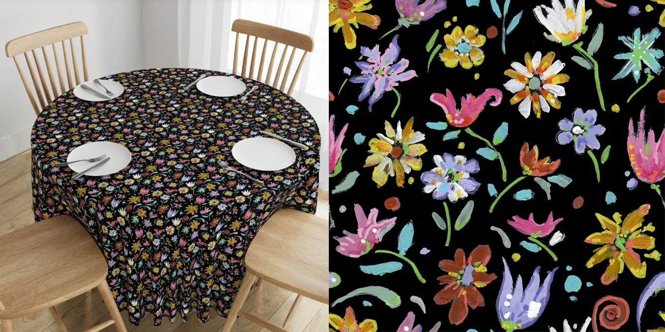 A round dining table covered with a black tablecloth featuring a colorful floral pattern. The table is set with four white plates, each with silverware, and surrounded by three wooden chairs. The image also shows a close-up of the fabric pattern, which consists of various brightly painted flowers on a black background.