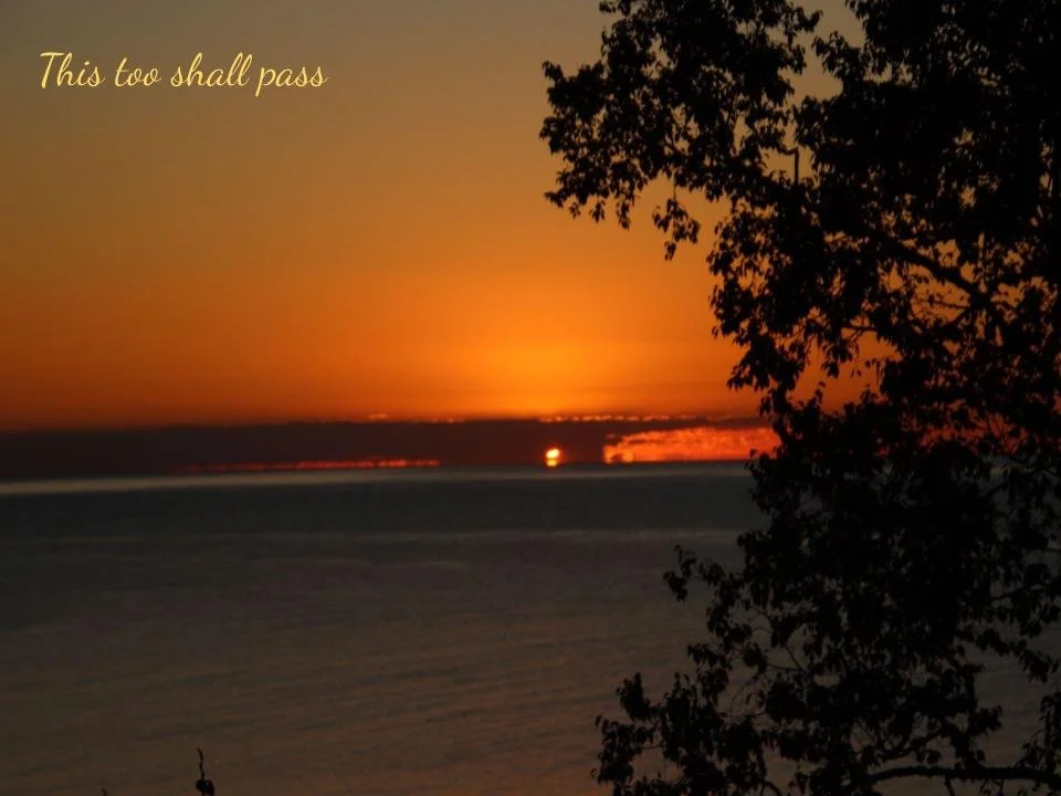 A sunset over a body of water with trees silhouetted on the right side and the phrase 'This too shall pass' written in yellow text in the upper left corner.