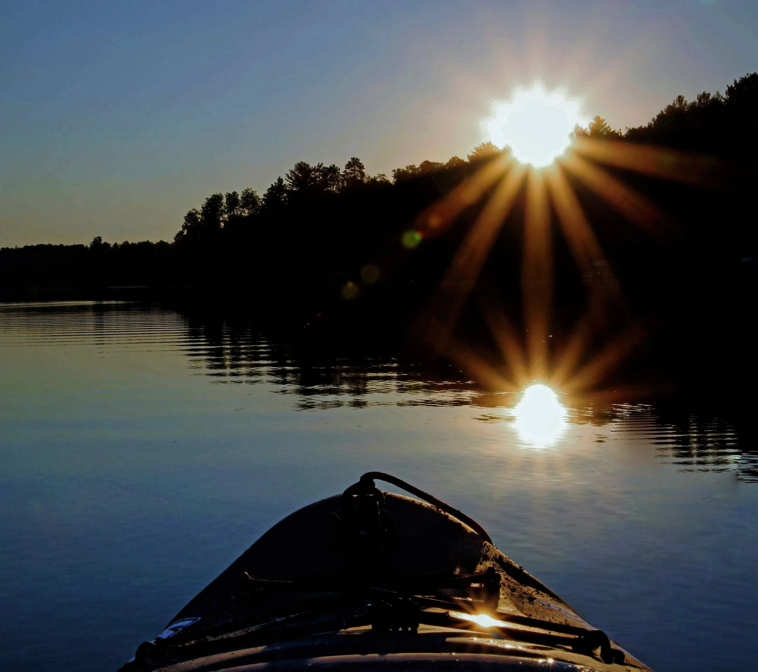 A boat on a calm lake at sunrise with the sun partially hidden behind a hill, creating sun rays and reflections on the water.