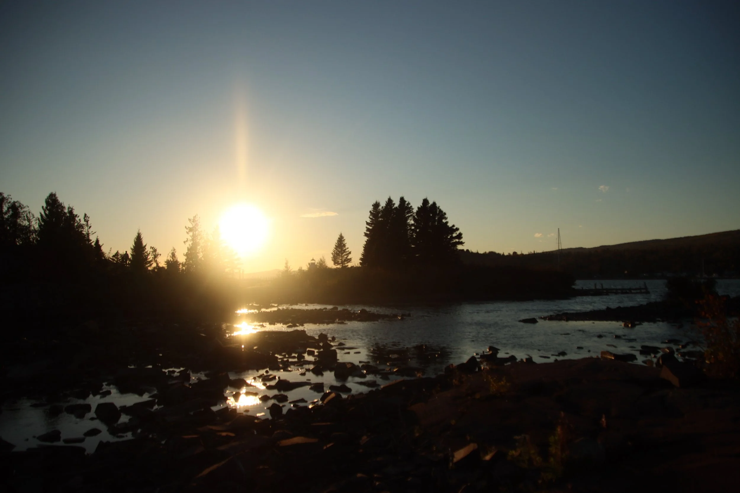 Sun setting over a river with trees silhouetted against the sky and rocks in the foreground.