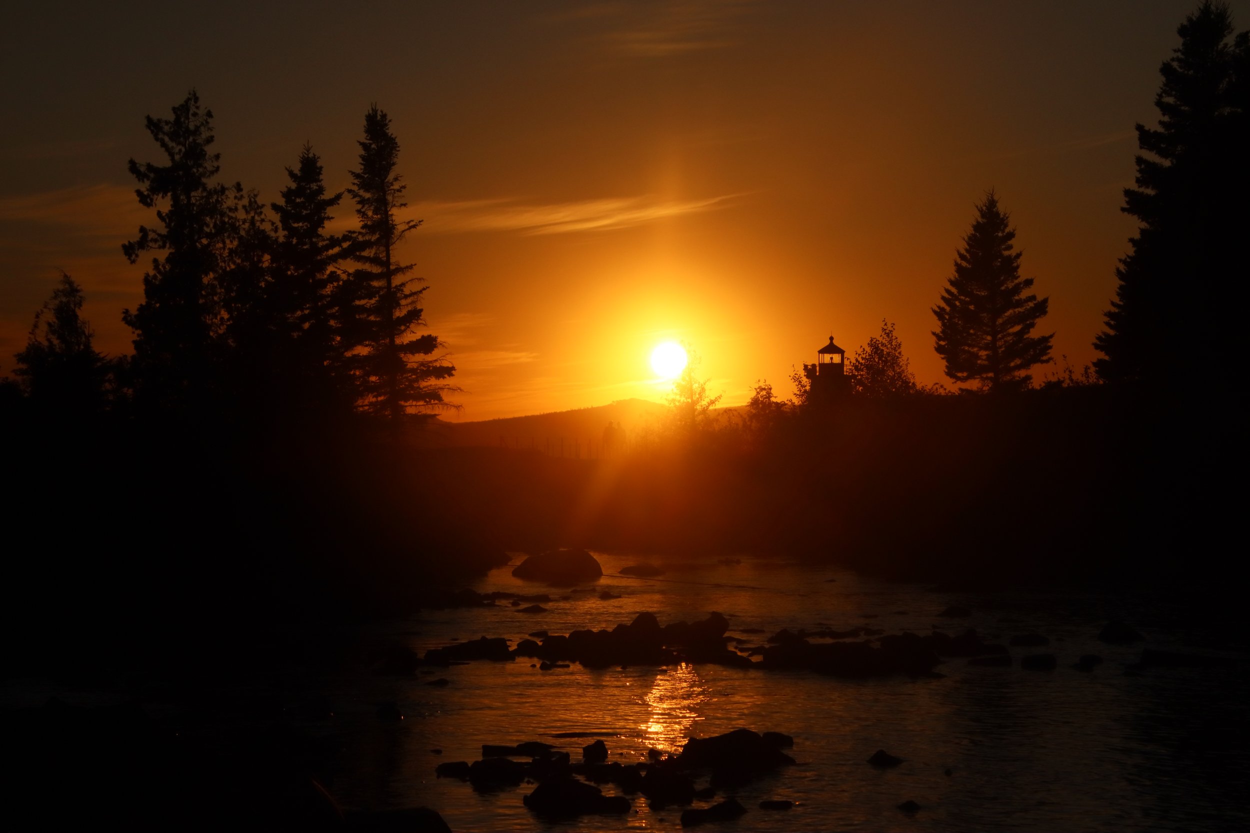 Sun setting behind trees and a small lighthouse, with a river and rocks in the foreground.