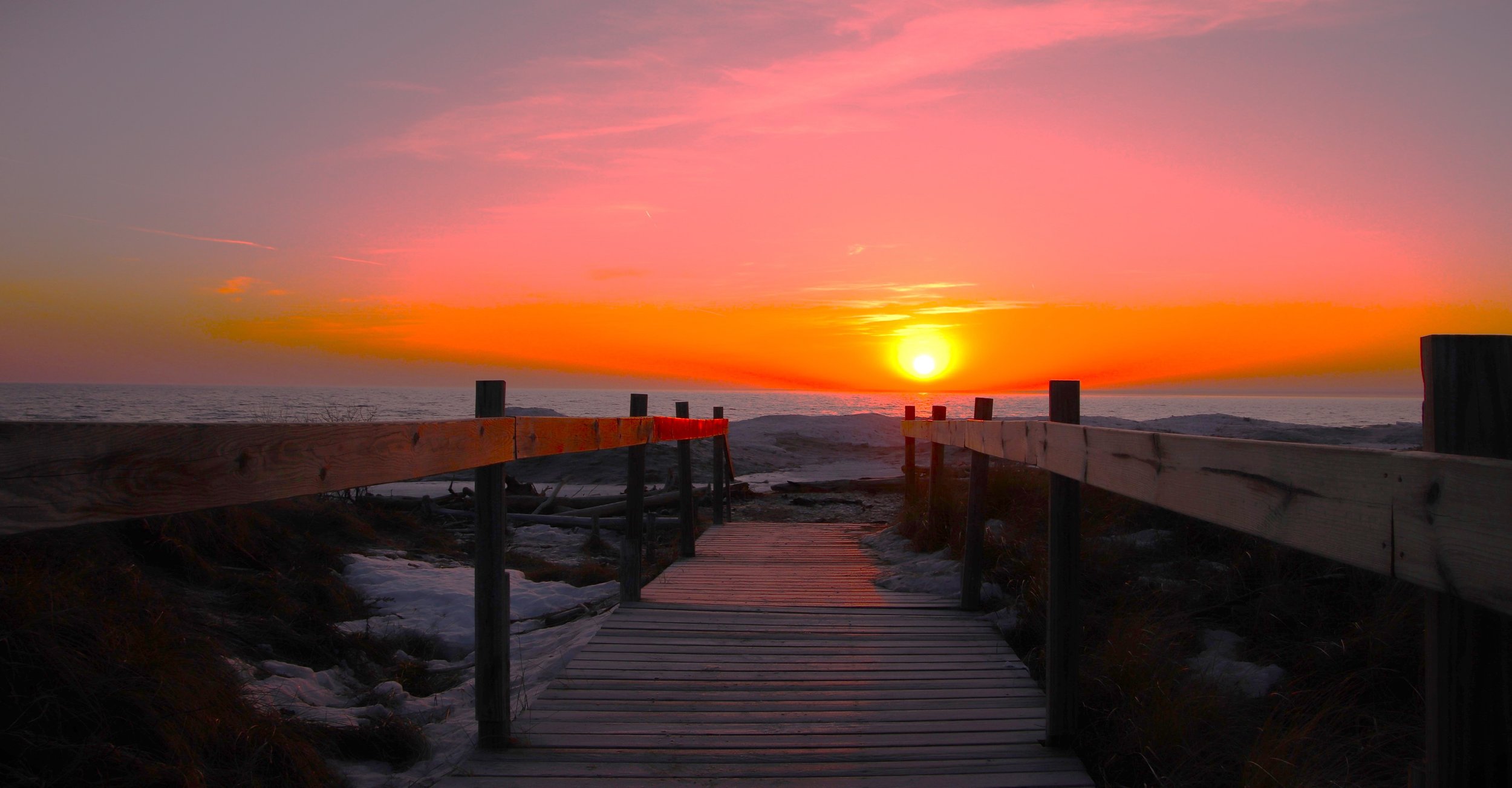 A sunset over the ocean viewed from a wooden boardwalk with snow on the ground.