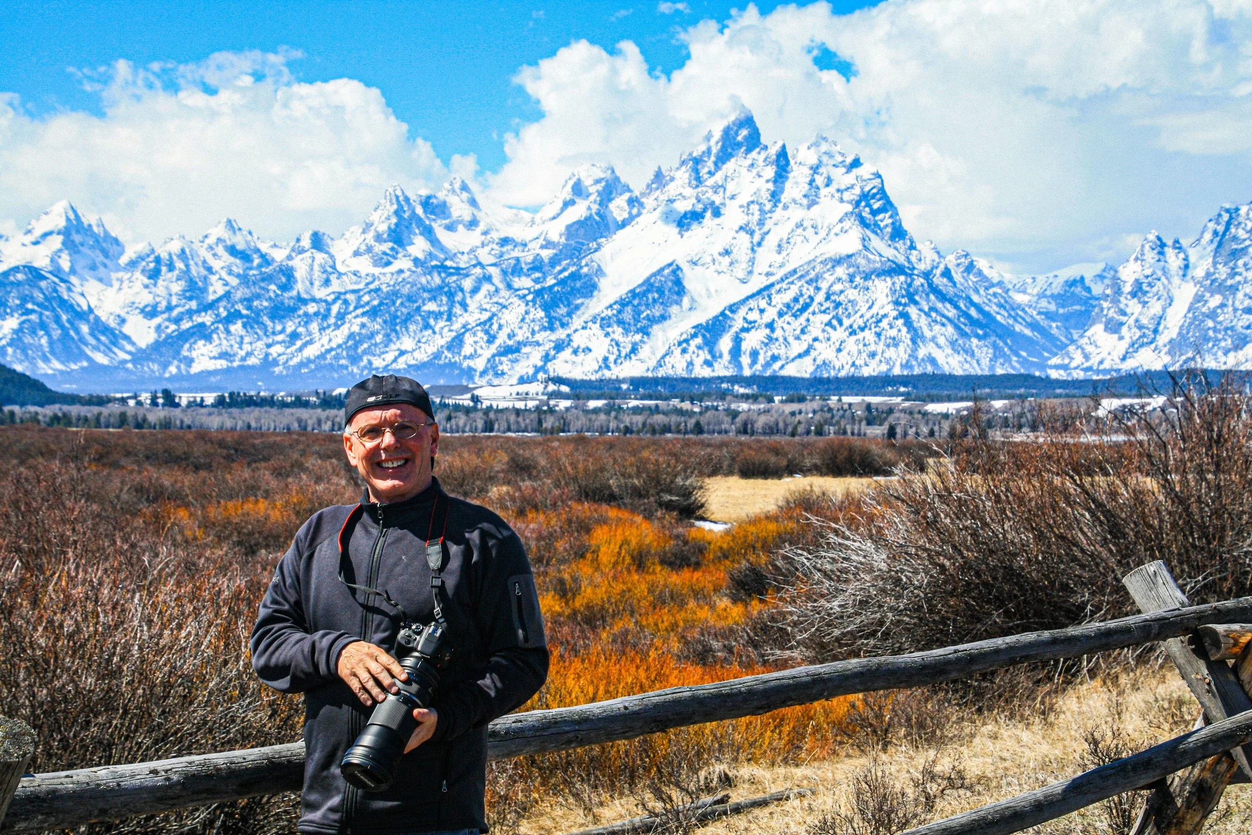 A man in outdoor gear, holding a camera, stands in front of a landscape with colorful autumn foliage, a fence, and snow-capped mountains under a partly cloudy blue sky.